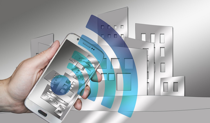 Hand hält ein Smartphone, aus dem ein blaues WLAN-Symbol hervorgeht; im Hintergrund stilisierte Gebäudegrafiken