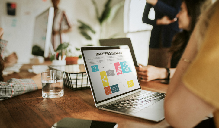 Laptop auf einem Holztisch mit einer geöffneten Marketing‑Strategie‑Grafik auf dem Bildschirm; im Hintergrund sitzen mehrere Personen an einem Tisch